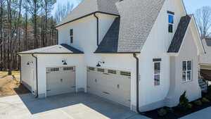 A two-story house with light-colored siding and dark shingles. It features a three-car garage with white doors and black handles. The driveway is concrete, and there are trees in the background.