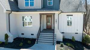 Front exterior of a modern house with a mix of white brick and vertical siding. It features a wooden door with glass panels, steps leading to the entrance, and small shrubs planted along the front.