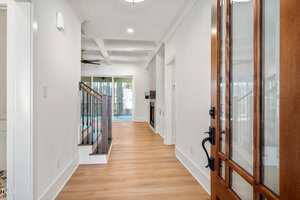 A hallway with light wooden flooring leads to a room with large glass doors. On the left, there are stairs with black metal railings. The walls are painted white, and a ceiling light fixture is visible.