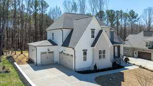 A two-story white house with a dark gray roof and a three-car garage. The driveway is concrete, and the house is surrounded by trees. There are no cars in the driveway, and neighboring houses are visible.