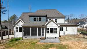 A two-story house with a grey roof and white siding is shown from the back. It features multiple windows, a screened porch, and a patio. The yard is mostly bare dirt with some grass. Other houses and trees are visible in the background under a clear blue sky.