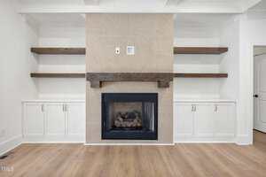 Modern living room with a central fireplace surrounded by beige tile. Dark wooden mantel above the fireplace, flanked by built-in wooden shelves. White cabinets with silver handles are below the shelves. Light wooden flooring and white walls.