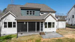 Rear view of a two-story house with a screened-in porch, gray siding, and a brown shingle roof. The backyard has a small grassy area and a paved section. Adjacent houses are partially visible. Clear blue sky in the background.