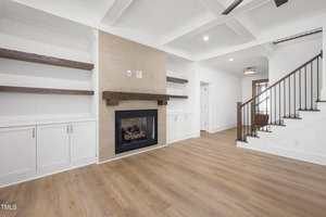 A modern living room features a built-in fireplace with wooden mantel, white shelves and cabinets on either side, white walls, and light wood flooring. A staircase with black railing is visible on the right.