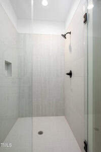 A modern, glass-enclosed shower with light beige tiles, a small niche on the wall, and a black showerhead. The space features a recessed ceiling light and a floor drain in the center.