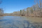 A calm river meanders past 0 Tilley Road, through a forested area with bare trees reaching under the clear blue sky. The distant bridge appears serene, while Bullock-like reflections of tree branches dance gracefully on the shimmering water's surface.