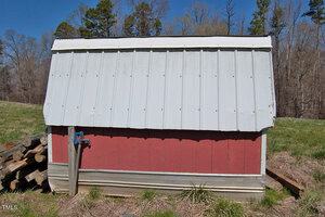 A small red structure with a white metal roof stands in a grassy area. A stack of firewood is visible to the left, and a blue faucet is attached to the front of the structure. Trees are in the background.