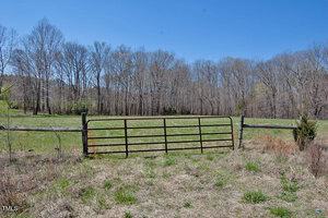 A rusty metal gate stands between two wooden posts, partially open in a field with dry grass. Beyond the gate is a green clearing and a forest of bare trees under a clear blue sky.