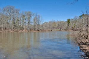 A calm lake is surrounded by leafless trees under a clear blue sky. The water's edge is visible in the foreground, and a small wooden bridge spans the lake in the distance. Sparse vegetation is present along the shoreline.
