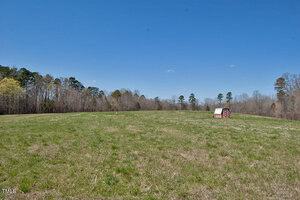 A wide, grassy field under a clear blue sky with a small red and white shed on the right side. The field is bordered by trees in the background.