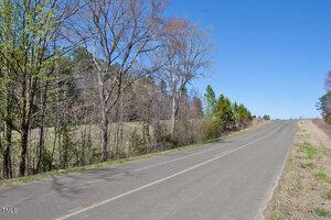 A paved road stretches into the distance under a clear blue sky. On the left, there are trees with some green leaves and bare branches. Grassy fields are visible on both sides of the road.