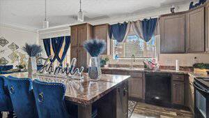 A modern kitchen features a central island with blue chairs and decorative items, including a "Family" sign. Wooden cabinets and a marble countertop contrast with dark appliances. A window with blue curtains provides natural light.