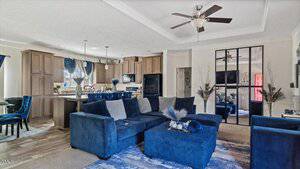 Modern living room with blue velvet sofas, a matching ottoman, and a light blue rug. A large mirror rests against a white wall, reflecting the kitchen area. Ceiling fans are on the white ceiling, and dark wood cabinets are seen in the kitchen area.