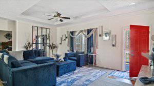 Living room with blue velvet sectional sofa and armchair, mirrors on walls, white curtains, and a light patterned rug. Ceiling fan overhead, decorative vases on a console table, and a red door ajar, letting sunlight in.