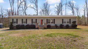 A single-story, gray mobile home on 830 Plum Nutty Road features a striking red front door, a small front porch, and black shutters. It's set in Henderson, surrounded by sparse grass and small bushes, with leafless trees silhouetted against a partly cloudy sky.