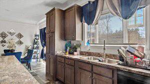 A kitchen with wooden cabinets and a granite countertop. The sink is equipped with a drying rack containing dishes. A window with curtains above the sink overlooks the outside. In the background, a living area with blue chairs and decorative items is visible.