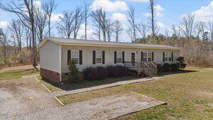 Single-story, light-colored manufactured home with a wooden front porch and steps. Located on a grassy lot with a gravel driveway. Surrounded by trees and a clear blue sky.