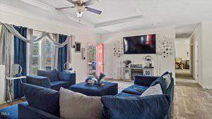 A living room features a large blue sectional sofa, a wall-mounted TV, and a ceiling fan. Blue and white curtains frame the windows. A console table beneath the TV displays decorative items, and a red door is slightly ajar, leading outside.