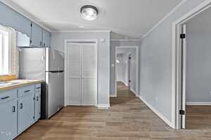 A kitchen area with light blue cabinets, a wooden countertop, and a stainless steel refrigerator. A hallway leads to other rooms. The floor is wood, and the walls are painted light blue with white trim. A ceiling light fixture is visible.