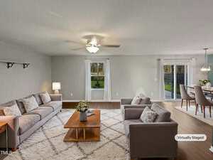 A virtually staged living room features two gray sofas, a wooden coffee table on a white rug, and a ceiling fan. A dining area is visible in the background with a table and chairs. Large windows offer a view of the backyard.