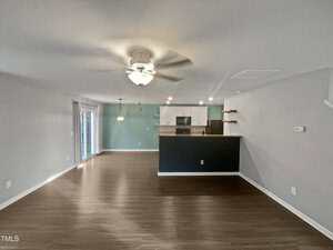 Open-plan living area with wood flooring, featuring a ceiling fan and a hanging light fixture. The room leads to a kitchen with a breakfast bar and stainless steel appliances. Sliding doors on the left open to a patio area. Walls are painted light gray.