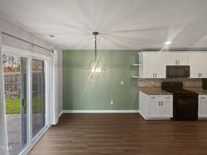 A modern kitchen and dining area with light wood flooring. It features white cabinets, a black stove, and a globe-shaped light fixture. The wall is painted green, and there is a sliding glass door leading to an outdoor yard.