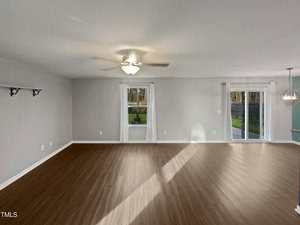 An empty room with light gray walls and wooden flooring. It features a ceiling fan with lights, two windows with white curtains, and a sliding glass door leading to an outdoor area. Shelves are mounted on one wall.