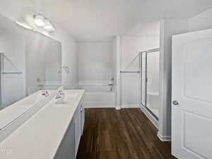 A modern bathroom featuring a dual sink vanity with a large mirror, white cabinetry, a bathtub, a glass-enclosed shower, and wood-style flooring. The space is bright with a wall-mounted light fixture above the mirror.