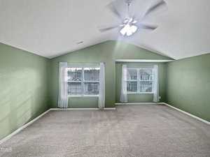 A spacious, carpeted room with light green walls and a vaulted ceiling is shown. It has a ceiling fan with a light, two windows with sheer white curtains, and an airy atmosphere. The windows overlook a suburban neighborhood.