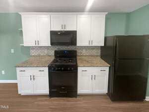 A kitchen with white cabinets, a black microwave, and a black gas stove with an oven. The countertops are granite, and there's a black refrigerator to the right. A gray and white tile backsplash is above the counter. The walls are painted green.