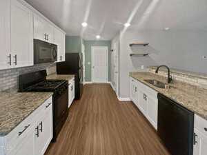 A modern kitchen with white cabinets, granite countertops, and wooden floors. Includes a black stove, refrigerator, and dishwasher. The walls are light gray, featuring open shelves above the sink, and a doorway near the kitchen's end.