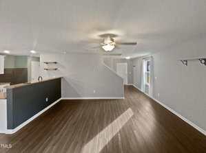 Spacious, empty room with dark wood flooring and light gray walls. A ceiling fan with lights is in the center. A partial view of a kitchen with open shelving is on the left, and a small window is on the right. A staircase leads to another level.