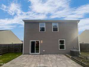 The image shows the back view of a two-story house with brown siding. There are two windows on the upper floor and a sliding glass door on the ground level, leading to a stone patio. The yard is fenced, and the sky is clear with some clouds.
