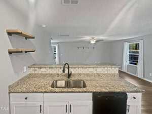 Open kitchen view with granite countertops, a dual sink, and black faucet. White cabinets and a subway tile backsplash line the wall. The kitchen overlooks a living area with a ceiling fan, wooden shelves, and a window with vertical blinds.