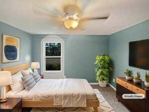 A bedroom with a double bed, two side tables with lamps, and a dresser with plants and a mounted TV. The walls are painted light blue, and there's a large window. The room features a ceiling fan and is labeled "Virtually Staged.