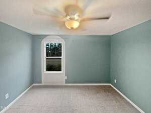 An empty room with light blue walls and beige carpet. A ceiling fan with a light is on, and a window on the left wall shows trees outside. The room is well-lit and has a minimalist appearance.