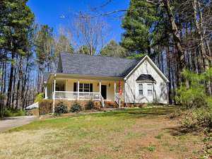 A single-story house with a gabled roof, a covered front porch, and a concrete driveway. The front yard is grass-covered and bordered by trees. A flag is displayed near the entrance. The sky is clear and blue.