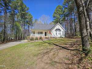 A single-story house with a gable roof and front porch, surrounded by trees. An American flag is displayed near the entrance. A driveway leads to the house, which is set on a grassy lawn under a clear blue sky.