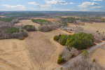 Aerial view of a rural landscape featuring expansive fields, clusters of trees, and a winding road. A small cluster of buildings is visible in the center. The sky is partly cloudy, and the terrain is a mix of brown and green hues.