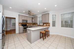 Modern kitchen with beige cabinets, stainless steel appliances, and a central island with a white countertop and two wooden stools. The floor is tiled, and there are three windows with blinds. Ceiling fan and recessed lighting are visible.