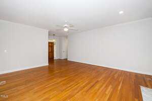 An empty room features polished hardwood floors, white walls, and a white ceiling with a ceiling fan and recessed lighting. A doorway leads to a hallway with a visible wooden door. No furniture is present.