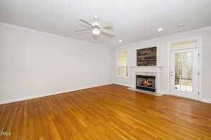 A spacious, empty living room with polished wooden floors. The room features a ceiling fan, white walls, recessed lighting, and a fireplace with a wooden mantel. Large windows and a glass door provide natural light.