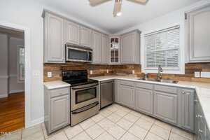 A kitchen with gray cabinets, stainless steel appliances, and a double-basin sink. The countertop is light-colored, and the backsplash has a wood-like texture. A window with blinds is above the sink, and the floor is tiled.