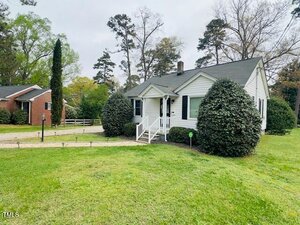 A single-story white house with a dark roof is surrounded by a well-maintained lawn. There are large bushes flanking the front entrance, which has white stairs and a railing. Trees and a neighboring house are visible in the background.