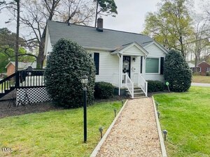 A small white house with a dark roof, surrounded by greenery, features a front porch and a pathway leading to the entrance. Bushes flank the porch, and a lamppost is alongside the walkway. Trees and additional houses are visible in the background.