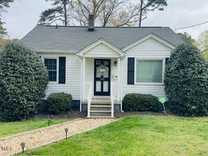 A small white house at 100 Pine Tree Drive features a dark roof and a central entrance flanked by two bushes. The door, adorned with a wreath, is accessible via a path leading to the steps. The lush green lawn boasts an Oxford ambiance, with trees framing the serene backdrop and a sign on the right.