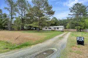 A mobile home on 4296 Tommie Sneed Road in rural Oxford features a gravel driveway. Tall trees and an open grassy field surround it, while a lawnmower rests near the road. The sky above is partly cloudy.