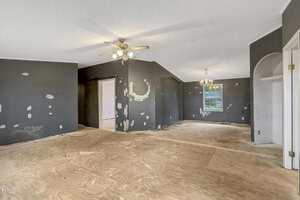 An empty room with unfinished plywood flooring and dark walls with visible patches. Ceiling fans with lights are on, and a window allows natural light in. A doorway leads to another room, and there is a chandelier hanging near the window.