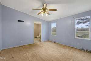 An empty room with light blue walls and a plywood floor. There is a ceiling fan with lights, two windows, and an open doorway leading to a small room with a visible toilet. A TV mounting bracket is on one wall.