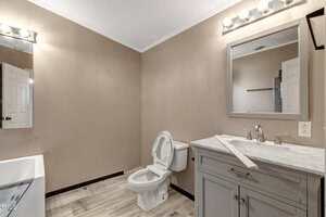 A bathroom with beige walls featuring a toilet, a vanity with a marble countertop, and a mirror. The floor is tiled, and a bathtub is partially visible on the left. The lighting consists of four bulbs above the mirror.